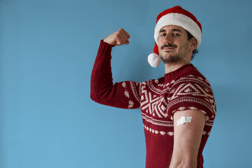 vaccine strong for Christmas. Confident smiling young man poses with santa claus hat and red sweater with arm bandage over blue background