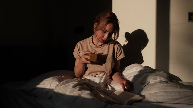 beautiful woman sitting in bed and reading a book in her pajamas with juice in her hand on sunny morning,woman in bed drinking juice and reading book on sunny morning changes page