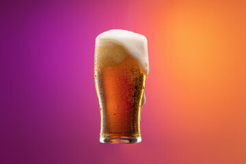 Full glass of frothy light lager beer isolated over gradient purple and orange color background in...