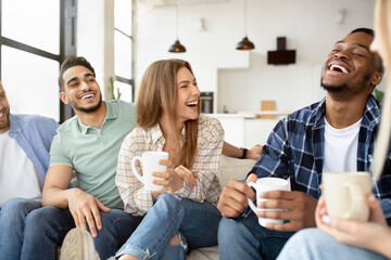 Group of happy multiracial friends drinking coffee in living room, having conversation, enjoying time together