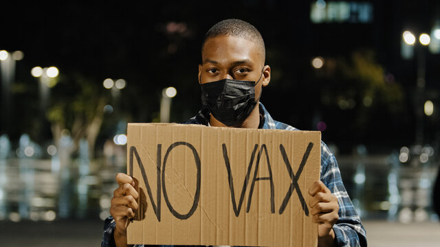 Young Angry African American Man In Black Mask Stands In City At Night Evening Holding Sign Banner With Text No VAX Afro Guy Protest Of Covid 19 Vaccine Stop Immunization Concept. Vaccination Phobia