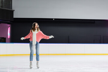 full length of astonished woman in white scarf, sweater and ear muffs skating on ice rink.