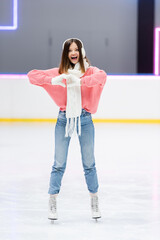 full length of cheerful woman in white scarf and ear muffs showing heart with hands while skating...