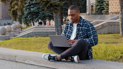 Fototapeta premium Afro american student guy african business man sitting outdoors in city street typing on laptop working online with computer e-learning listening to music on headphones singing song chatting in net