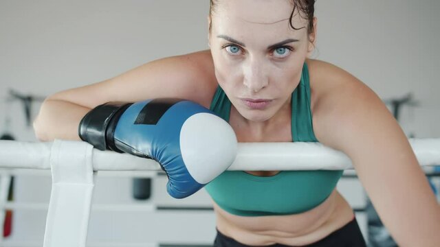 Slow motion portrait of tired young female boxer wearing gloves resting in boxing ring corner breathing hard and looking at camera. Sports and people concept.