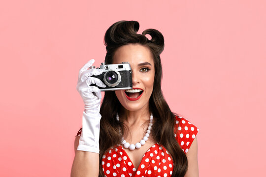 Portrait of young pinup female with retro camera taking pictures over pink studio background