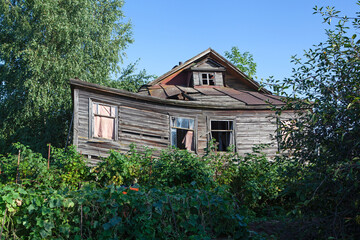 Fototapeta na wymiar Old abandoned rustic wooden house is surrounded by the greenery of cherry orchard. Rural area. Crumbling building