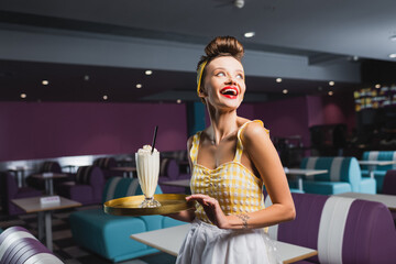 cheerful pin up waitress with tattoo holding tray with milkshake in cafe.