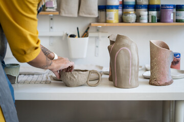 Working table of craft ceramist with ceramics tableware. Female artist shaping potter kitchenware to bake in studio. Woman in apron creating crockery in creative studio. Self-employed pottery business