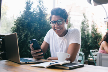 Portrait of cheerful software developer in optical eyewear for vision protection holding cellphone technology in hand and smiling at camera during time for freelance programming via digital netbook