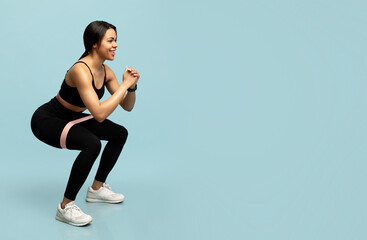 Training for glute muscles. Sporty black woman doing deep squat exercises with elastic bands, blue...