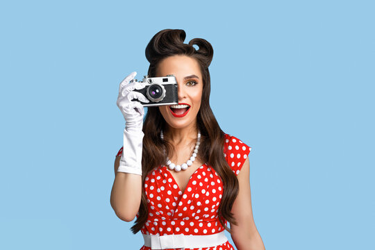 Portrait of cheerful pinup woman in polka dot dress taking photos with retro camera on blue studio background