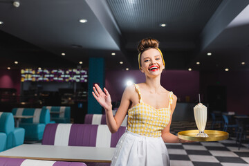 excited young pin up waitress holding tray with milkshake and gesturing in cafe.