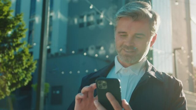 Slow motion attractive businessman walking near office center uses phone texting scrolling tapping smile. Outdoors sunny. Feel happy. Portrait.