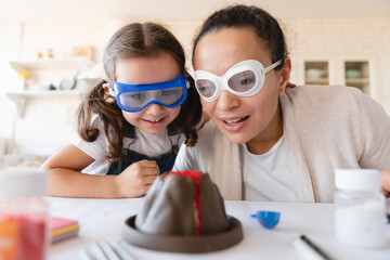 Closeup cropped shot of mother and daughter in protective eyewear watching volcano baked soda eruption at home kitchen together, making school science project. Home chemistry lab - obrazy, fototapety, plakaty