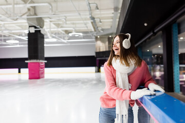 excited woman in ear muffs, scarf and pink sweater on ice rink.