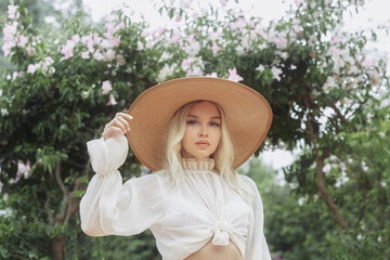 Portrait of romantic stylishly dressed young adult beautiful woman relaxing in a countryside park looking away. Blonde Parisian-styled model in a white transparent dress and straw hat standing against