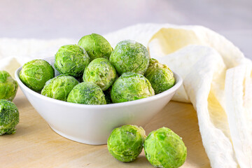 Many frozen whole brussels sprouts in white bowl on light wooden background.
