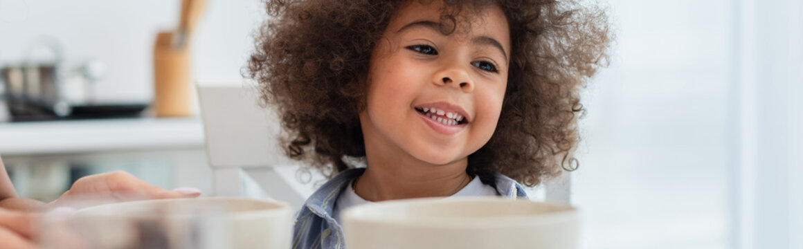 Smiling african american kid sitting near bowls and mother in kitchen, banner.
