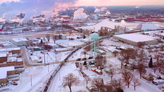 Long freight train slowly rolls through snow covered industrial area, aerial view.
