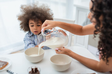 Blurred african american mother pouring milk in bowls near daughter in kitchen.