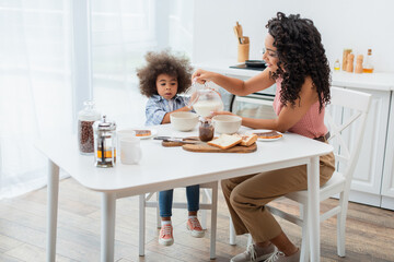 African american mom pouring milk near breakfast and daughter at home.