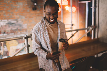 Joyful hipster guy in stylish spectacles using cellular gadget for network socialising during free time, happy African American man in messaging via application on smartphone device and smiling