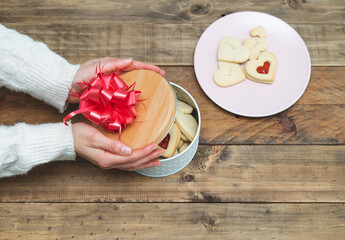 Valentine's Day. Woman's hands uncovering biscuit box with red ribbon and heart-shaped biscuits. Copy space.