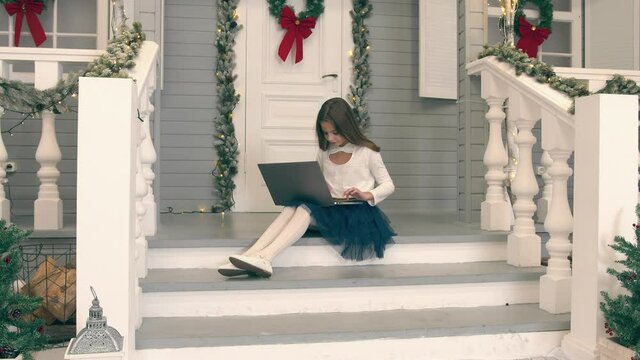 Child making a video call using a laptop. The girl sits on the doorstep of her house and communicates via video link holding a laptop on her lap