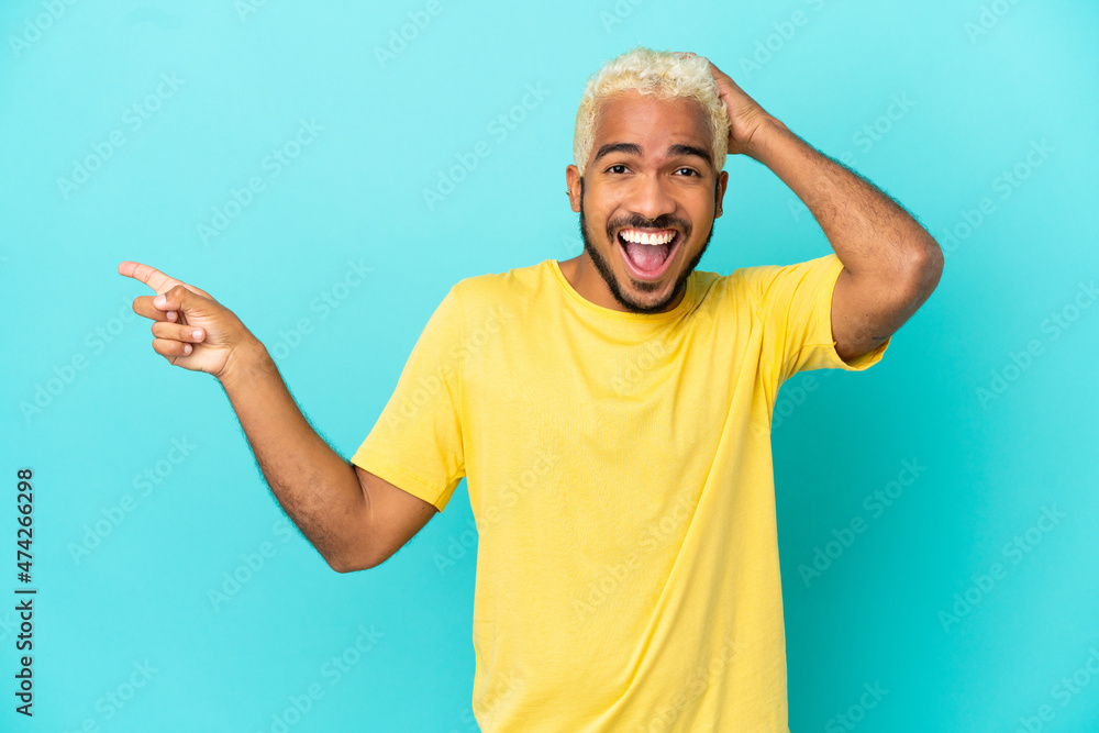 Sticker Young Colombian handsome man isolated on blue background surprised and pointing finger to the side