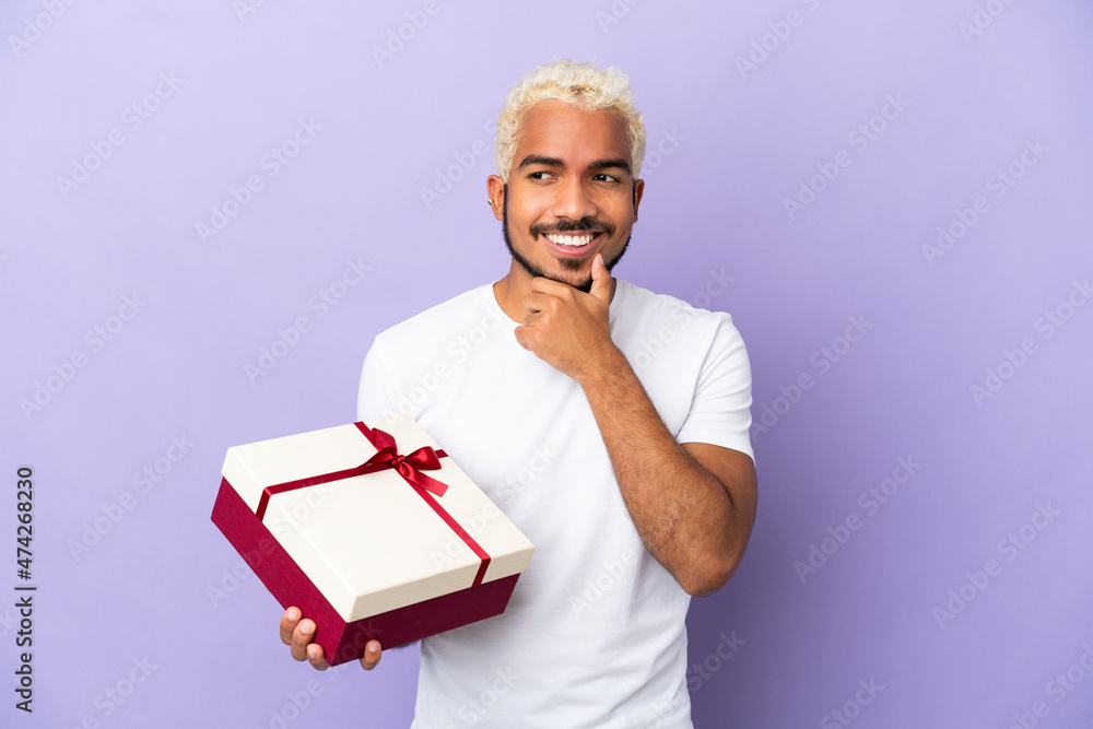 Poster young colombian man holding a gift isolated on purple background looking to the side and smiling