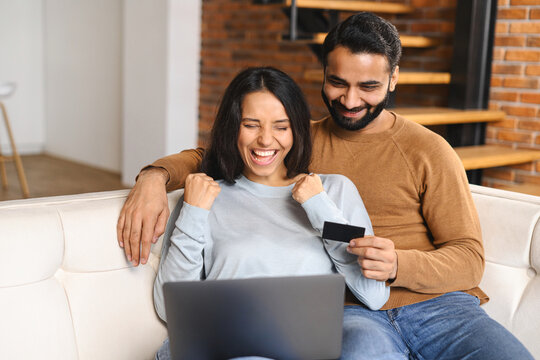 Cheerful Indian couple watching at laptop screen and smiling toothy. Overjoyed woman rejoicing while having shopping online with her husband. Transferring money, online banking concept