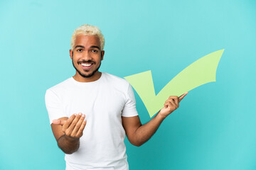 Young Colombian handsome man isolated on blue background holding a check icon and doing coming gesture
