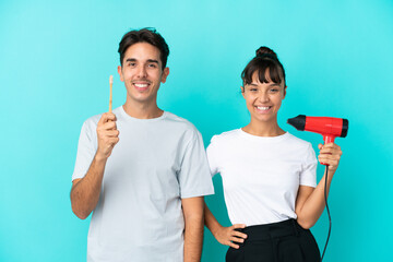 Young mixed race couple holding a hairdryer and toothbrush isolated on blue background posing with arms at hip and smiling