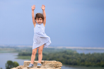 A little girl with her hands raised up on top of a mountain on a blurry background of a picturesque...
