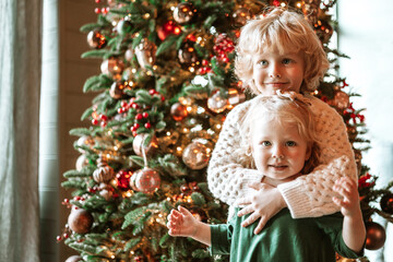 siblings children boy and little girl with blond hair and blue eyes in beautiful clothes are standing in living room near Christmas tree and decorating it family celebration of Christmas and New Year
