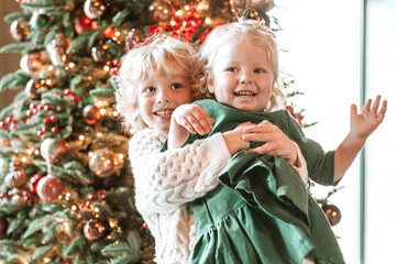 siblings children boy and little girl with blond hair and blue eyes in beautiful clothes are standing in living room near Christmas tree and decorating it family celebration of Christmas and New Year