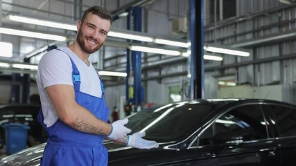 Happy technician young brunet mechanic man 20s wears denim overalls white t-shirt gloves work in vehicle repair shop point hands on car show thumb up like gesture on light workshop background indoors