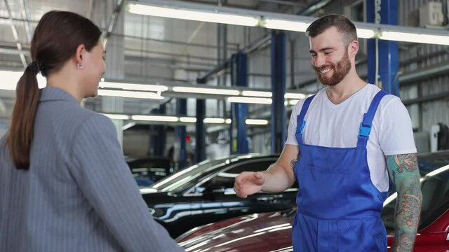 Young brunet mechanic man 20s wears denim overalls white t-shirt work in vehicle repair shop communicate pass car key to business woman customer handshake on light modern workshop background indoors