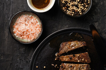 A piece of cooked beef meat steak on black plate cut in slices, white and black peer, coarse sea salt
