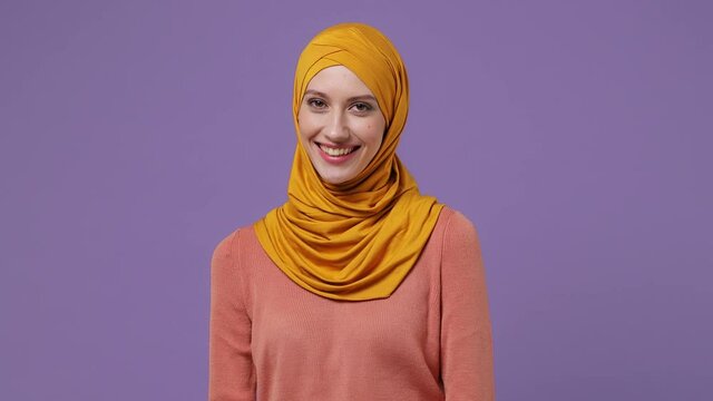 Smiling young arabian asian muslim woman in abaya hijab yellow clothes look camera wink eye blink isolated on plain pastel light violet background studio portrait. People uae islam religious concept