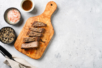 A piece of cooked beef meat steak on wooden board cut in slices, white and black peer, coarse sea salt. Top view, negative space