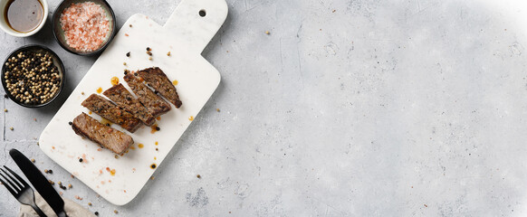 A piece of cooked beef meat steak on white marble board cut in slices, white and black peer, coarse sea salt. Top view, negative space, banner