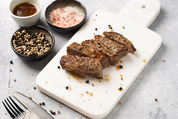 A piece of cooked beef meat steak on white marble board cut in slices, white and black peer, coarse sea salt