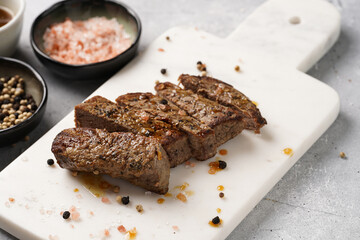 A piece of cooked beef meat steak on white marble board cut in slices, white and black peer, coarse sea salt