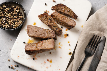 A piece of cooked beef meat steak on white marble board cut in slices, white and black peer, coarse sea salt