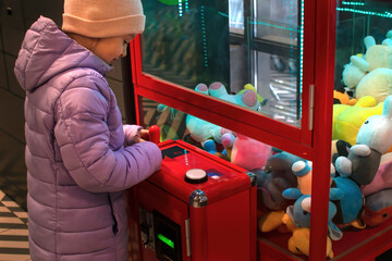 A child girl stands near a machine with soft toys and plays, wants to catch and pull out a toy prize