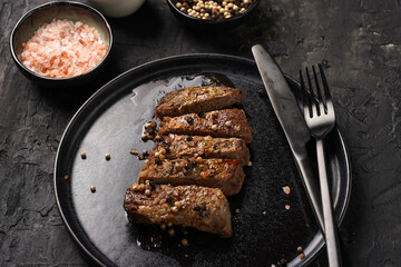 A piece of cooked beef meat steak on black plate cut in slices, white and black peer, coarse sea salt