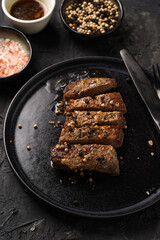 A piece of cooked beef meat steak on black plate cut in slices, white and black peer, coarse sea salt