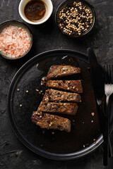 A piece of cooked beef meat steak on black plate cut in slices, white and black peer, coarse sea salt